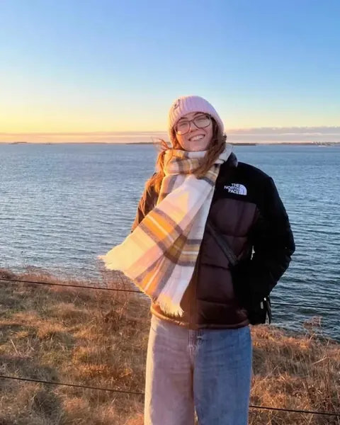 Grace in a hat and scarf, smiling by the water at sunset, with the fjord and a golden sky behind her.